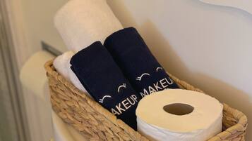 Un panier en osier contenant un rouleau de papier toilette blanc, deux serviettes bleu marine ornées de l’inscription « MAKEUP MAKEUP », et un objet argenté.