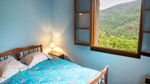 A bedroom with a bed, a nightstand with a lamp, and a window with a view of a forested landscape.