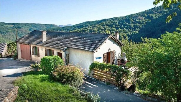 A two-story house with a tiled roof, surrounded by greenery and hills.