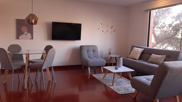Een moderne woonkamer met een glazen tafel, grijze stoelen, een grijze bank en een flatscreen televisie aan de muur.