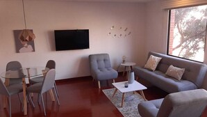 Een moderne woonkamer met een glazen eettafel, grijze banken en een flatscreen televisie aan de muur.