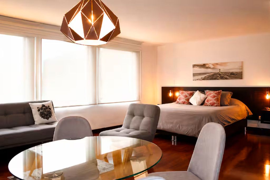 Habitación de hotel moderna con una mesa de vidrio, un sofá, una cama con almohadones decorativos y un ventanal grande.