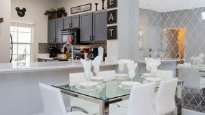 A modern kitchen with grey cabinets, a glass island, and a dining area set for six with white chairs and glassware.