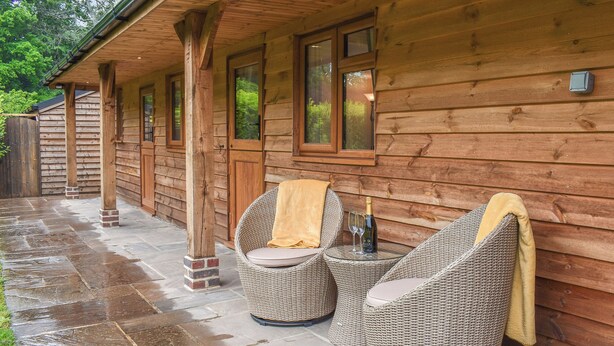A wooden cabin with a covered patio, wicker chairs, and a bottle of champagne. - The Stables at Early Autumn (Sevenoaks)