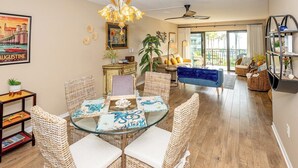 A spacious living room with a glass dining table, wicker chairs, a blue sofa, and a balcony with a view. - Follow The Sun at Ocean House 211 (St. Augustine)