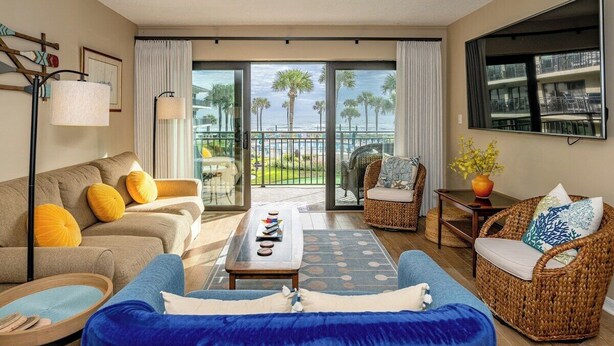 A spacious living room with a sofa, armchair, and a coffee table. There is a balcony with a view of palm trees and a beach. - Follow The Sun at Ocean House 211 (St. Augustine)