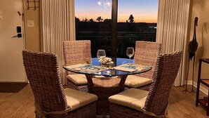 A dining area with a glass table surrounded by wicker chairs, a chandelier, and a view of the sunset through large windows. - Follow The Sun at Ocean House 211 (St. Augustine)