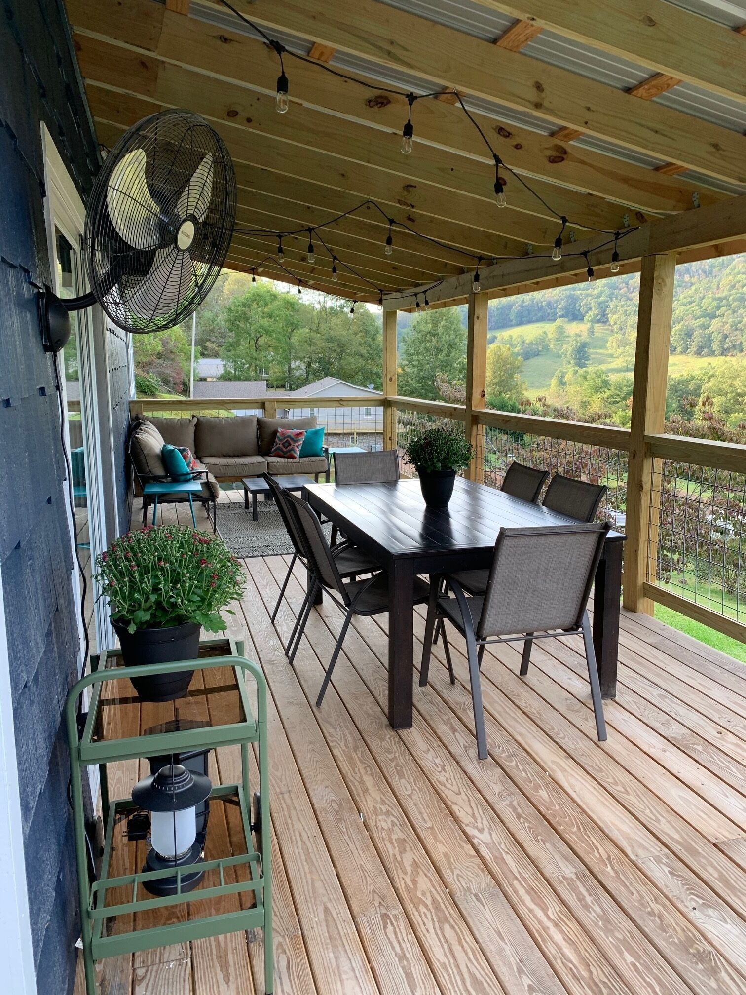 A covered outdoor patio with a wooden deck, a dining table with chairs, a sofa, and a fan.