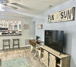 Cocina moderna con microondas, horno y pileta integrados. Hay una zona de comedor con mesa de madera, sillas y televisor. En la pared, un reloj y una cita motivacional.