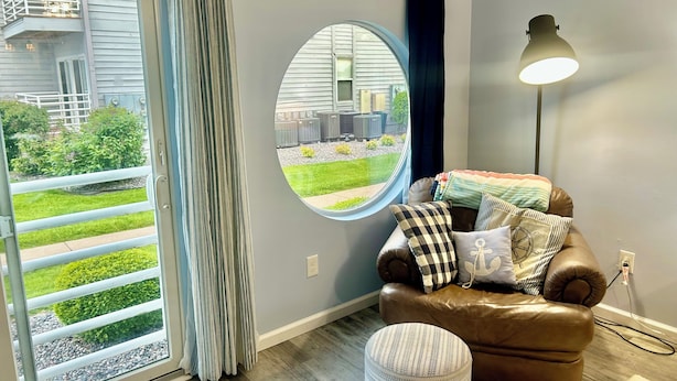 A room with a brown leather sofa, a round window, a floor lamp, and a view of a balcony with a railing.