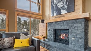 Una sala de estar acogedora con chimenea de piedra, sofá de cuero con cojines y amplias ventanas que ofrecen vistas a árboles cubiertos de nieve.
