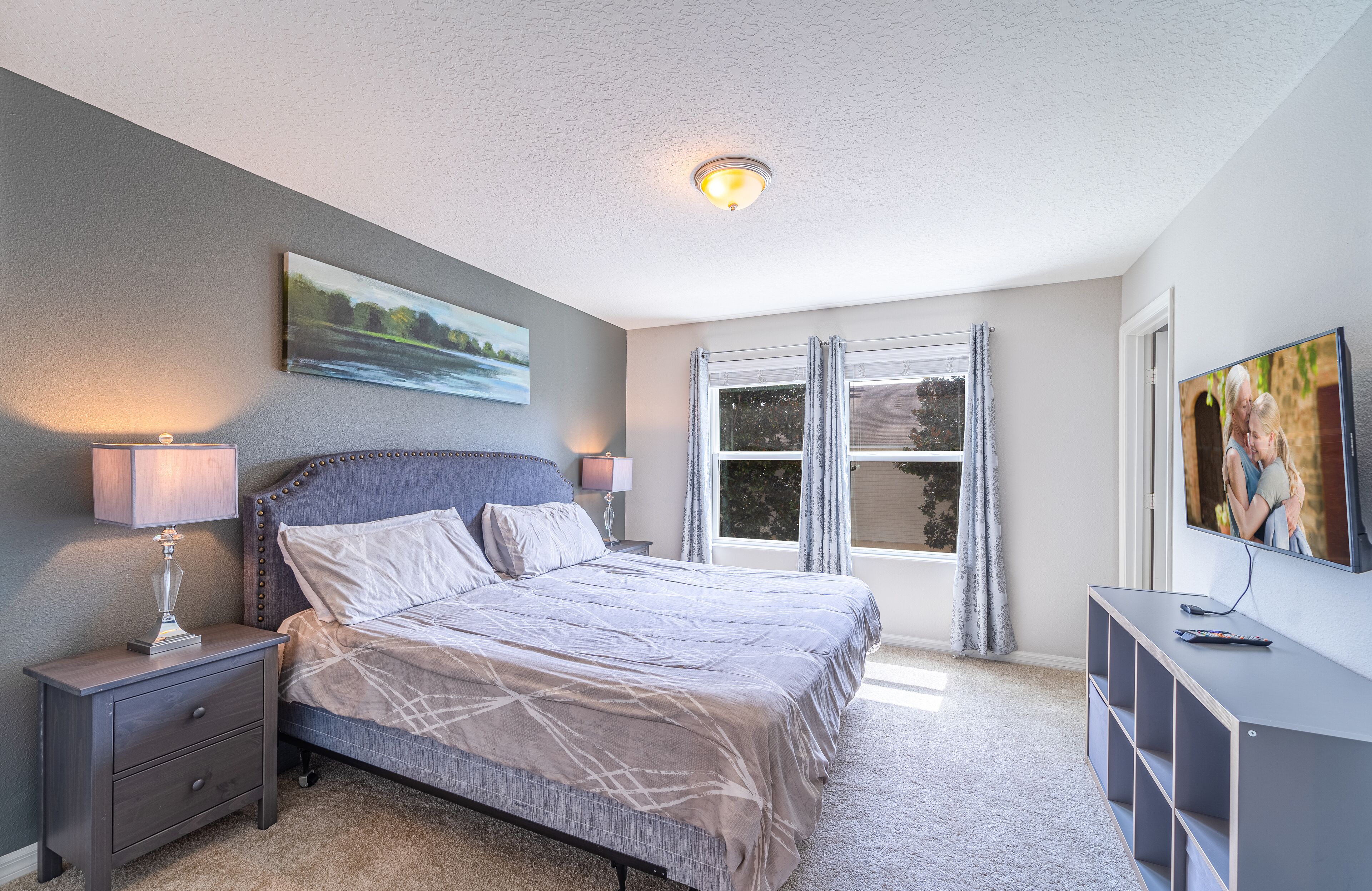 A bedroom with a bed, nightstand, lamp, TV, and a window with curtains.