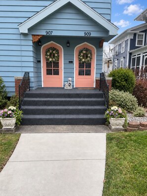 Une maison à deux étages, peinte en bleu, portant les numéros 907 et 909, avec un porche à l’avant doté de marches et orné d’une petite statue de chien.