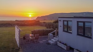 Vista al mar con atardecer, un pequeño edificio con balcón y un área verde con mesa y sillas.