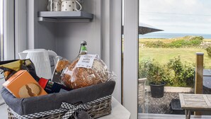Una cesta de mimbre con pan, una botella de vino y una cafetera sobre una mesa junto a una ventana con vistas a un jardín.