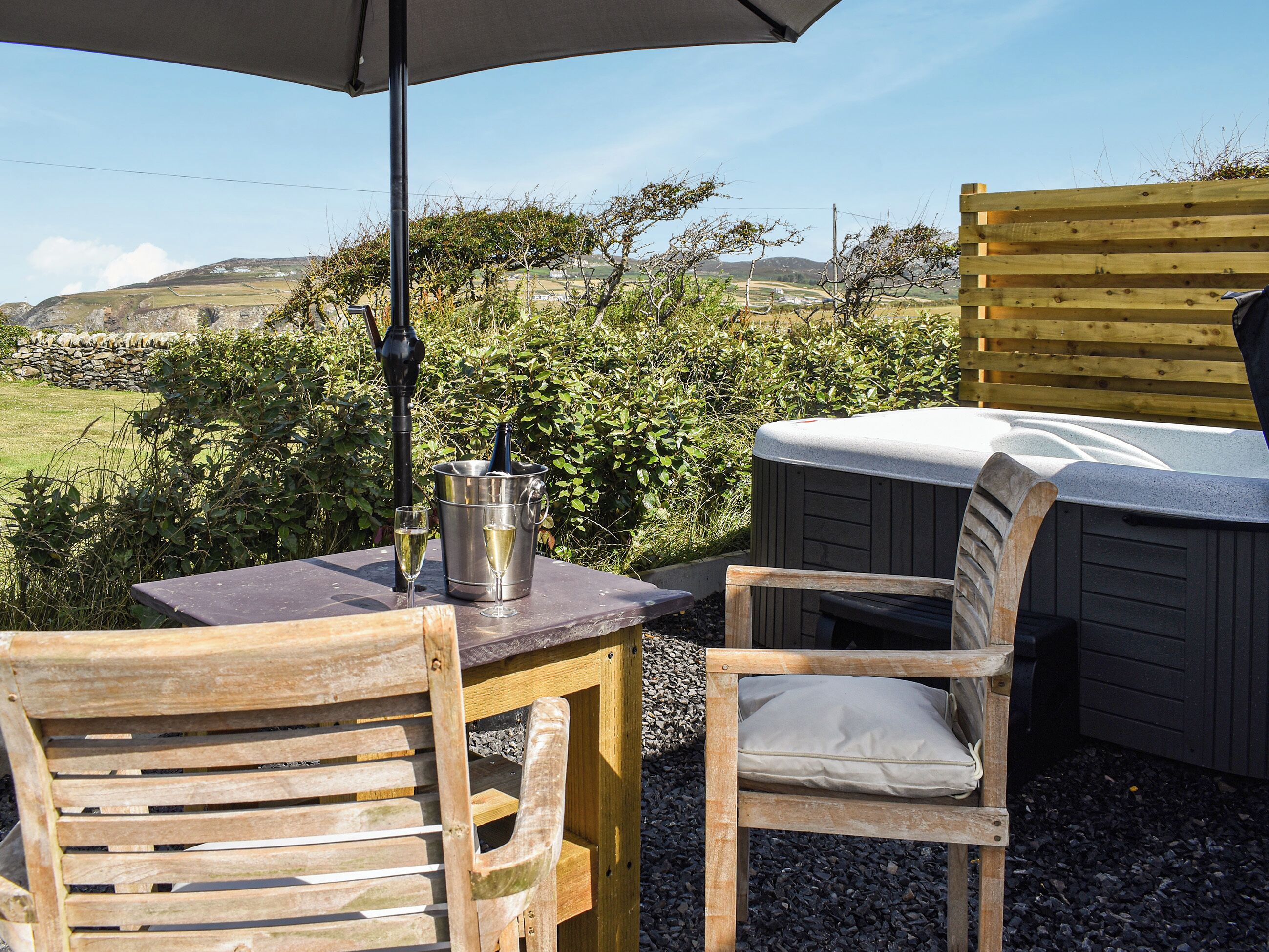 A table with a metal bucket, two glasses, and a bottle, set outdoors with a wooden chair and an umbrella.