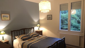 Un dormitorio con cama, lámparas de noche, ventana con vista a la vegetación y un radiador.