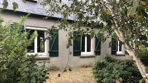 Una casa con un pequeño jardín, una ventana con contraventanas y un árbol en primer plano.