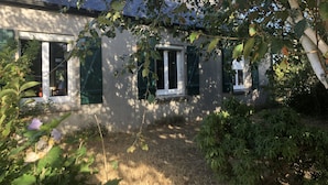 Una casa con techo inclinado, contraventanas verdes y un jardín con diversas plantas y un árbol pequeño.