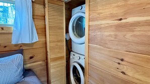 Una habitación de cabaña de madera, compacta, con cama, lavadora y ventana con cortinas.