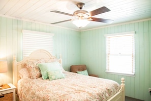 Une chambre à coucher avec un lit, une table de chevet, une chaise, un ventilateur de plafond et une fenêtre.