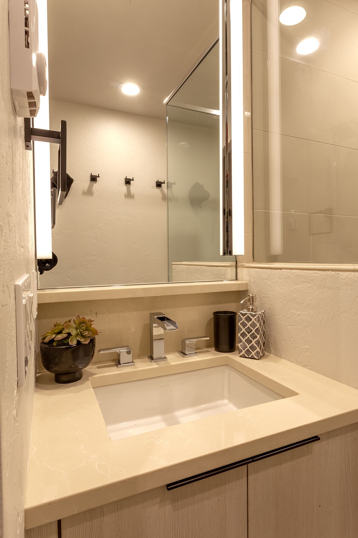 A bathroom with a large mirror, a sink, a faucet, a soap dispenser, and a small potted plant.