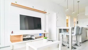 Ein modernes Wohnzimmer mit Flachbildfernseher, einem weißen Couchtisch und einer Essecke mit Barhocker.