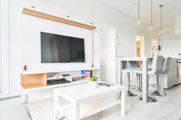 Una sala de estar moderna con un televisor de pantalla plana, una mesa de centro blanca y un comedor con taburetes de bar.