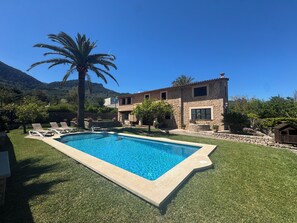 Une piscine bordée de pierres, un palmier et un bâtiment en pierre surmonté d’un toit en tuiles.