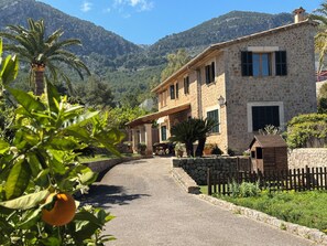 Un bâtiment en pierre, avec une route pavée menant à l’entrée, entouré de verdure et de montagnes.