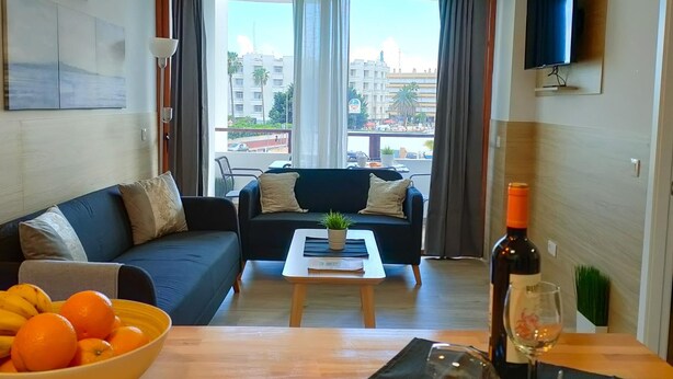 A modern living room with a sofa, a coffee table, and a TV. There is a balcony with a view of buildings and palm trees. - Stylish Friendly Holiday Home (Playa del Inglés)