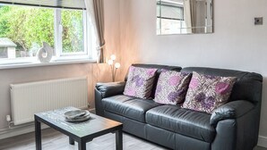 A modern living room with a black leather sofa, a small wooden table, and a window with curtains. - 1 bedroom accommodation in Bridlington (Bridlington)