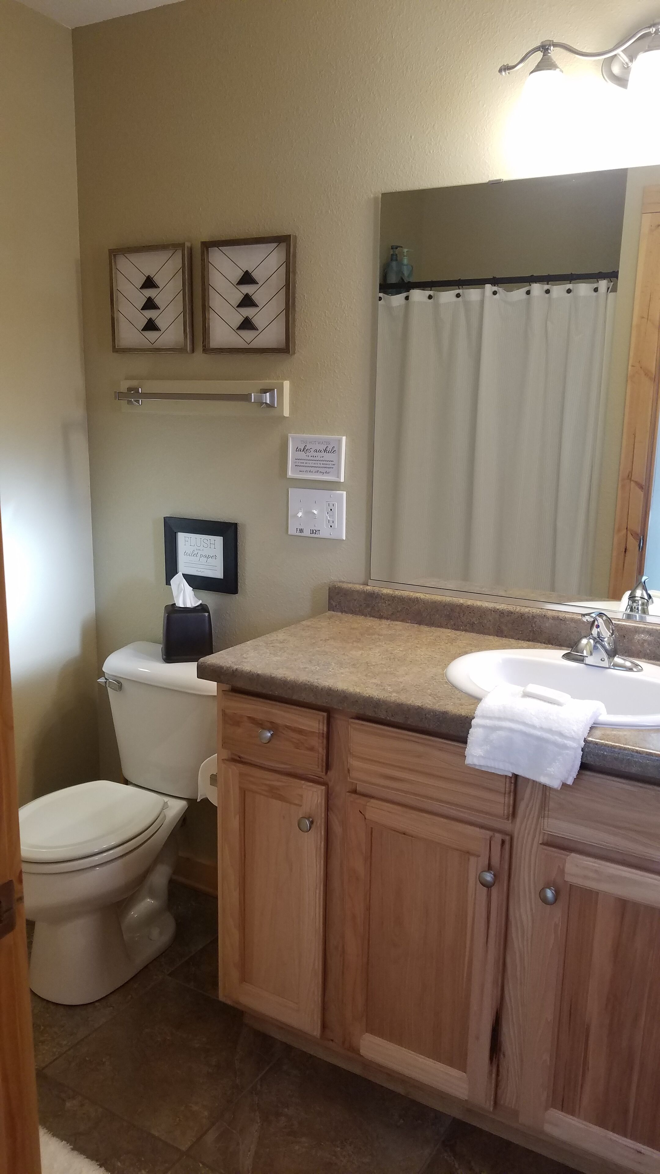 Baño con inodoro, lavabo y espejo. Hay dos cuadros enmarcados en la pared, un perchero y un cartel sobre el inodoro.