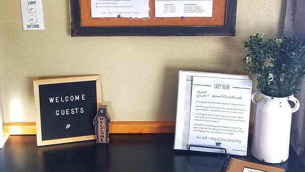 A desk with a corkboard displaying notices, a welcome sign, a guest book, and a potted plant.