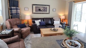 A cozy living room with a brown leather sofa, a wooden coffee table, and a fireplace. There are two lamps on either side of the sofa, and a framed picture of a bear on the wall.
