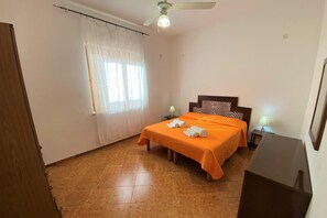 Un dormitorio con una cama, dos mesitas de noche con lámparas, un ventilador de techo y una ventana con cortinas traslúcidas.
