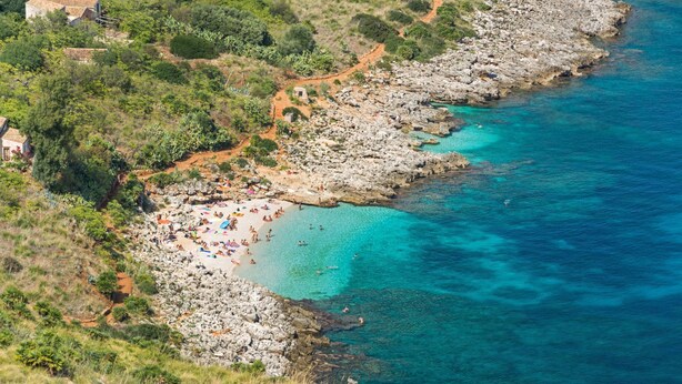 Zona costera con terreno rocoso, una playa con gente y un mar azul cristalino.