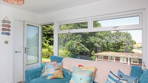 Ein heller Raum mit einem großen Fenster, einem blauen Sofa mit bunten Kissen und einer Korbschwinge als Lampe.