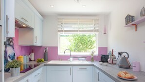 Eine moderne Küche mit weißen Schränken, einem großen Fenster mit Jalousien, einer Spüle und einer Arbeitsplatte mit Wasserkocher und einem Teller Brot.
