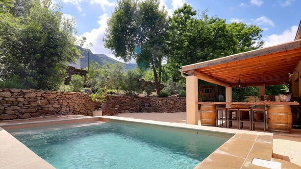 Una piscina con paredes de piedra, una pérgola de madera y un área con bancos de piedra.