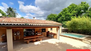 Un espacio al aire libre techado con una zona de barbacoa, mesas de picnic y una piscina.
