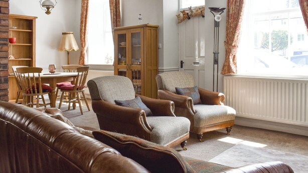 A cozy living room with a brown leather sofa, two armchairs, a wooden dining table with chairs, a cabinet, and a window with curtains.