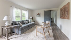 Un salon spacieux avec un canapé gris, une chaise en bois et une table avec des lampes. Une grande fenêtre laisse entrer la lumière naturelle, et une carte du monde est accrochée au mur.
