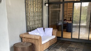 A wicker sofa with white cushions, a round wooden stool, and a glass door leading to an outdoor area with a view of a building and trees. - Beautiful and spacious loft in the Heart of Atlanta! (Atlanta)