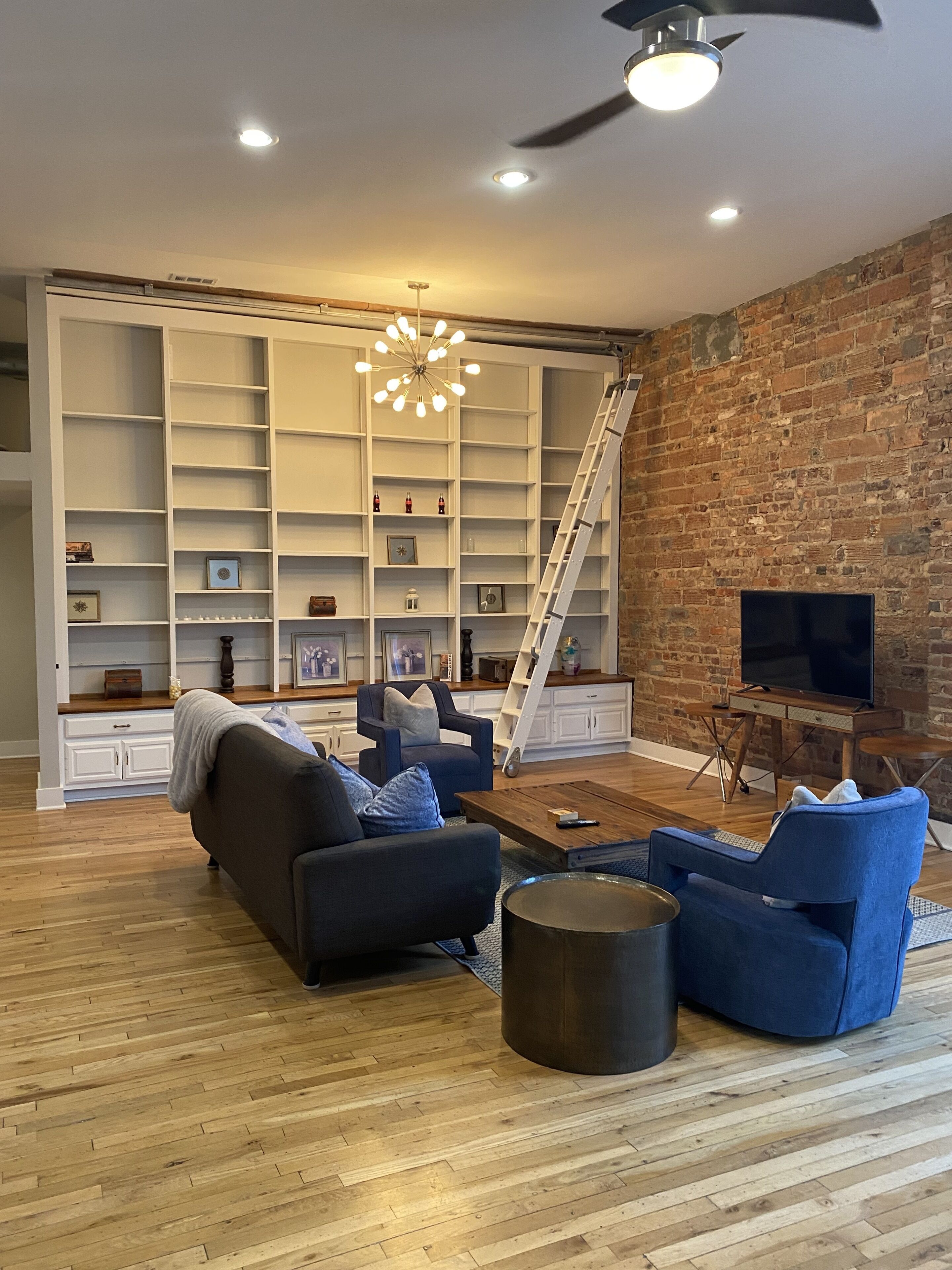 A modern living room with a brick wall, a white shelving unit, a black sofa, a coffee table, and a TV.