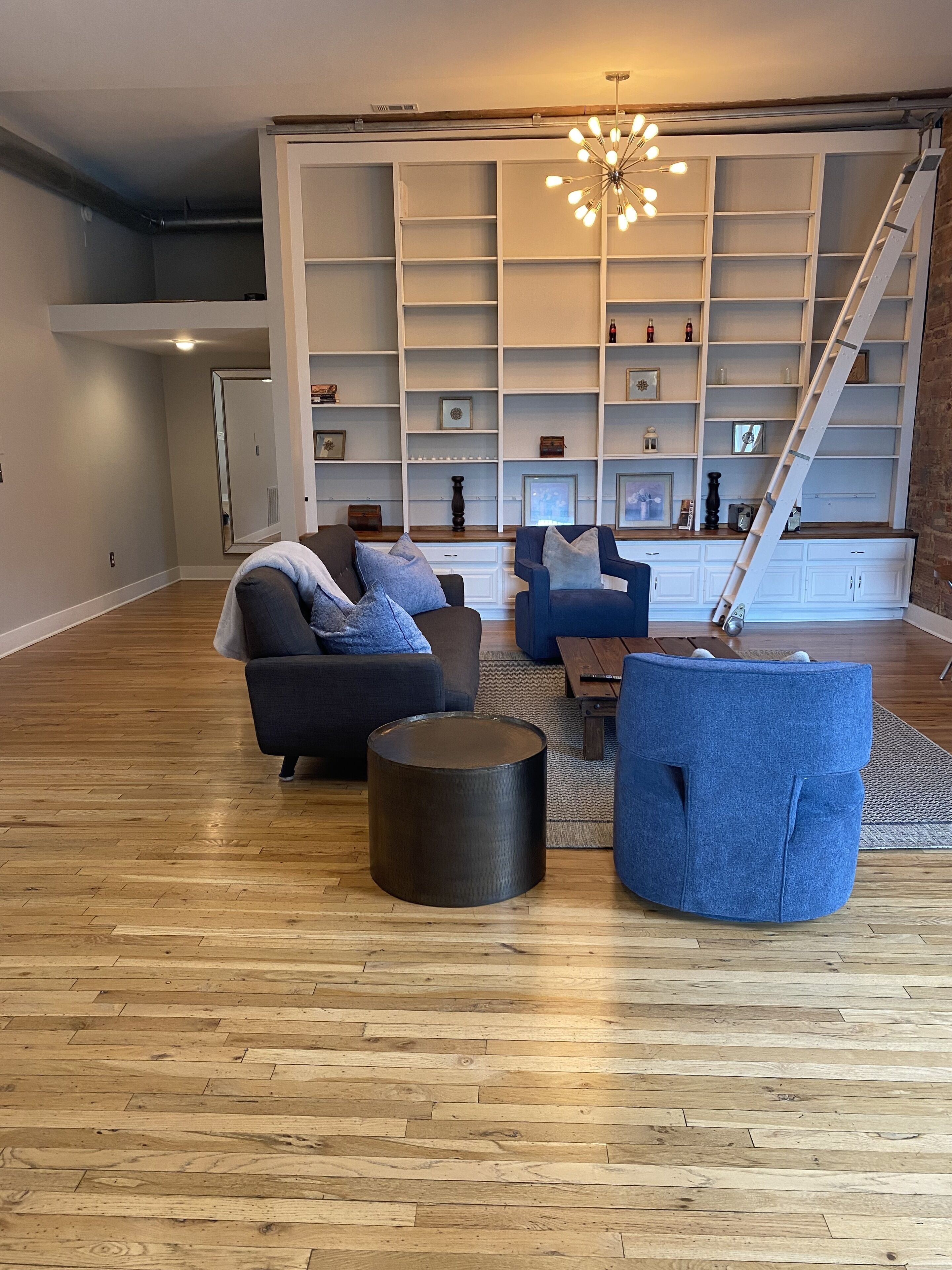 A spacious living room with a wooden floor, a large white shelving unit, and a modern chandelier.