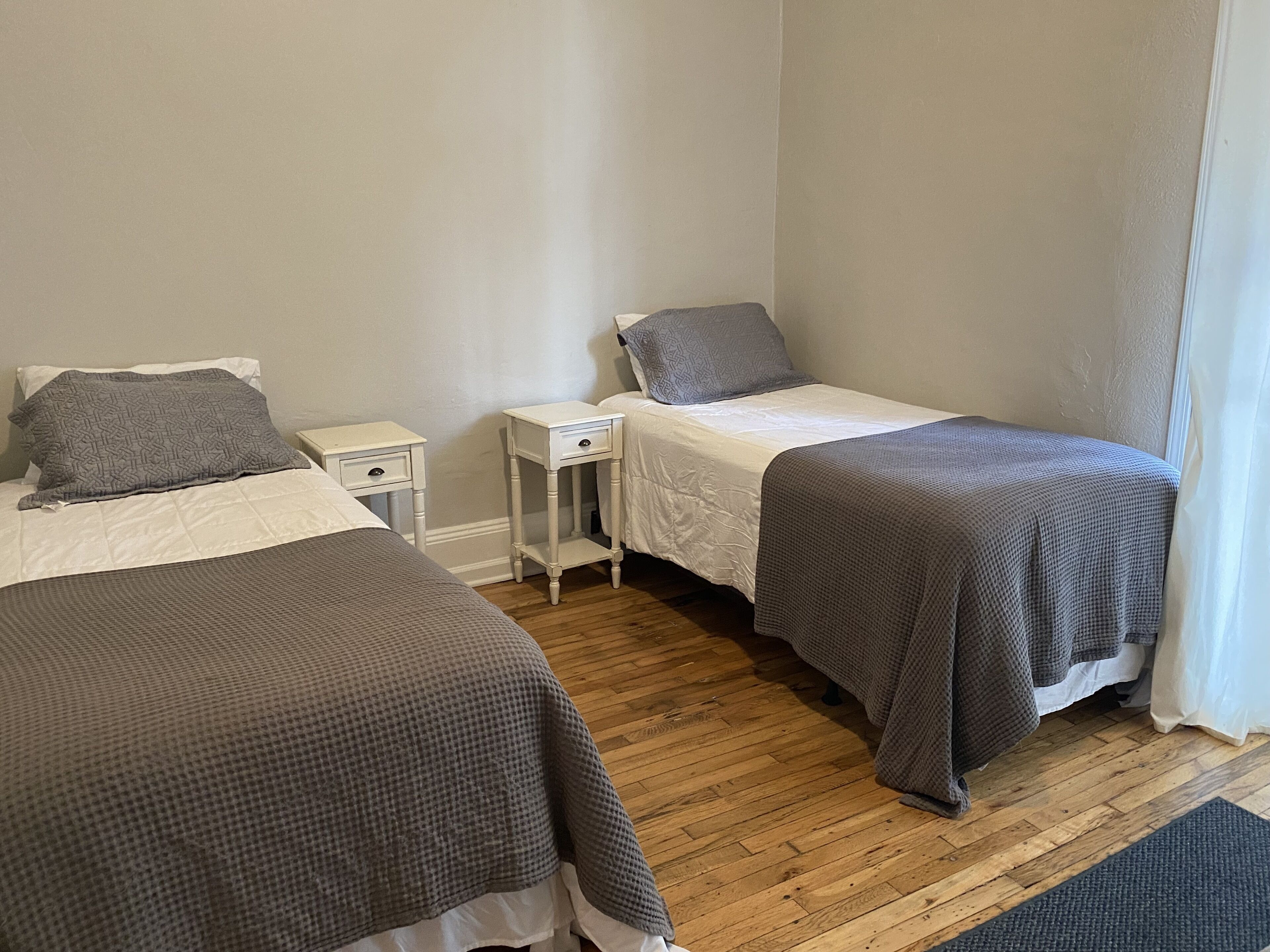 A room with two single beds, each with a pillow and a nightstand. The room has wooden flooring and white walls.