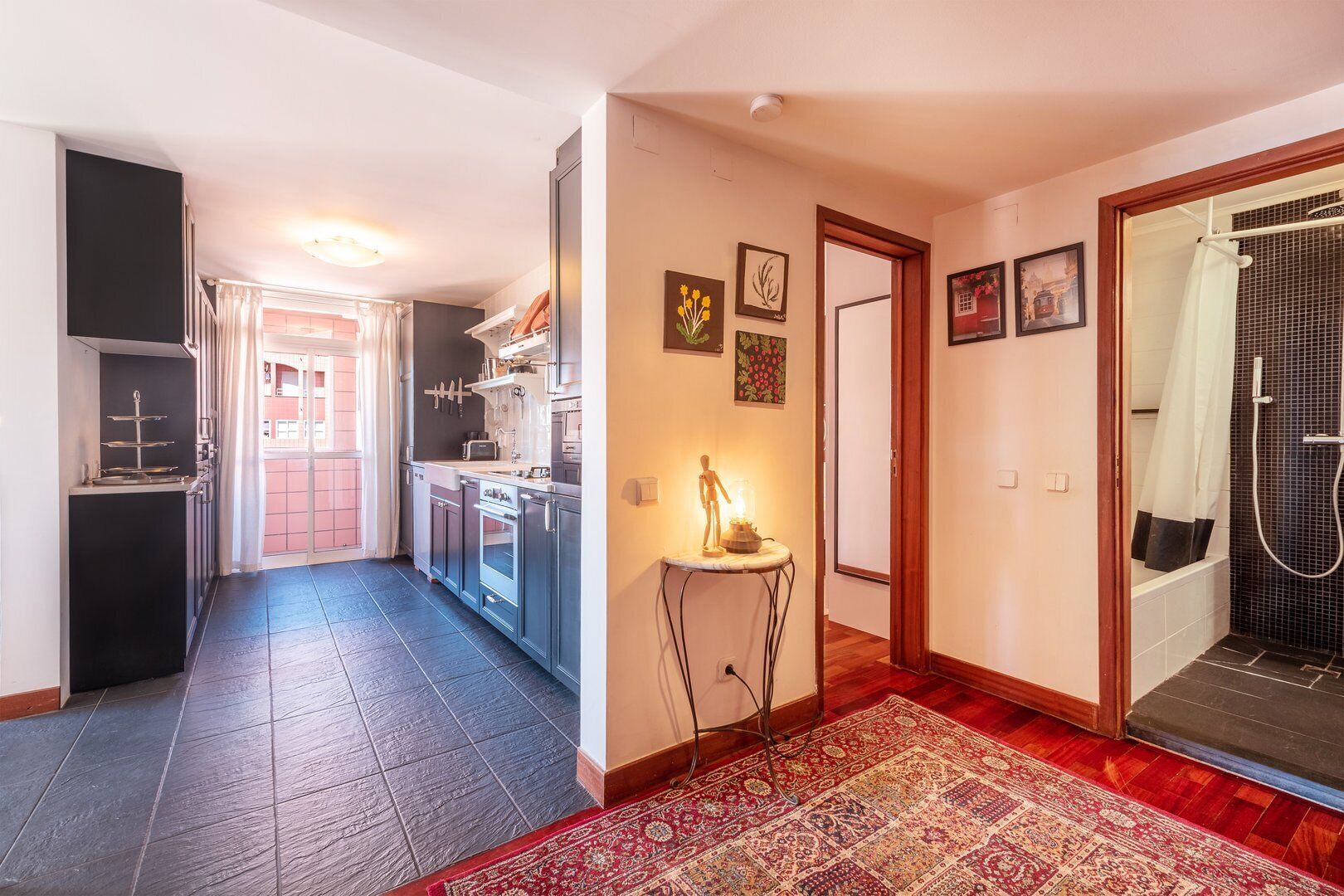 A hallway leading to a kitchen with a dining area, a small table with a lamp, and a shower area.