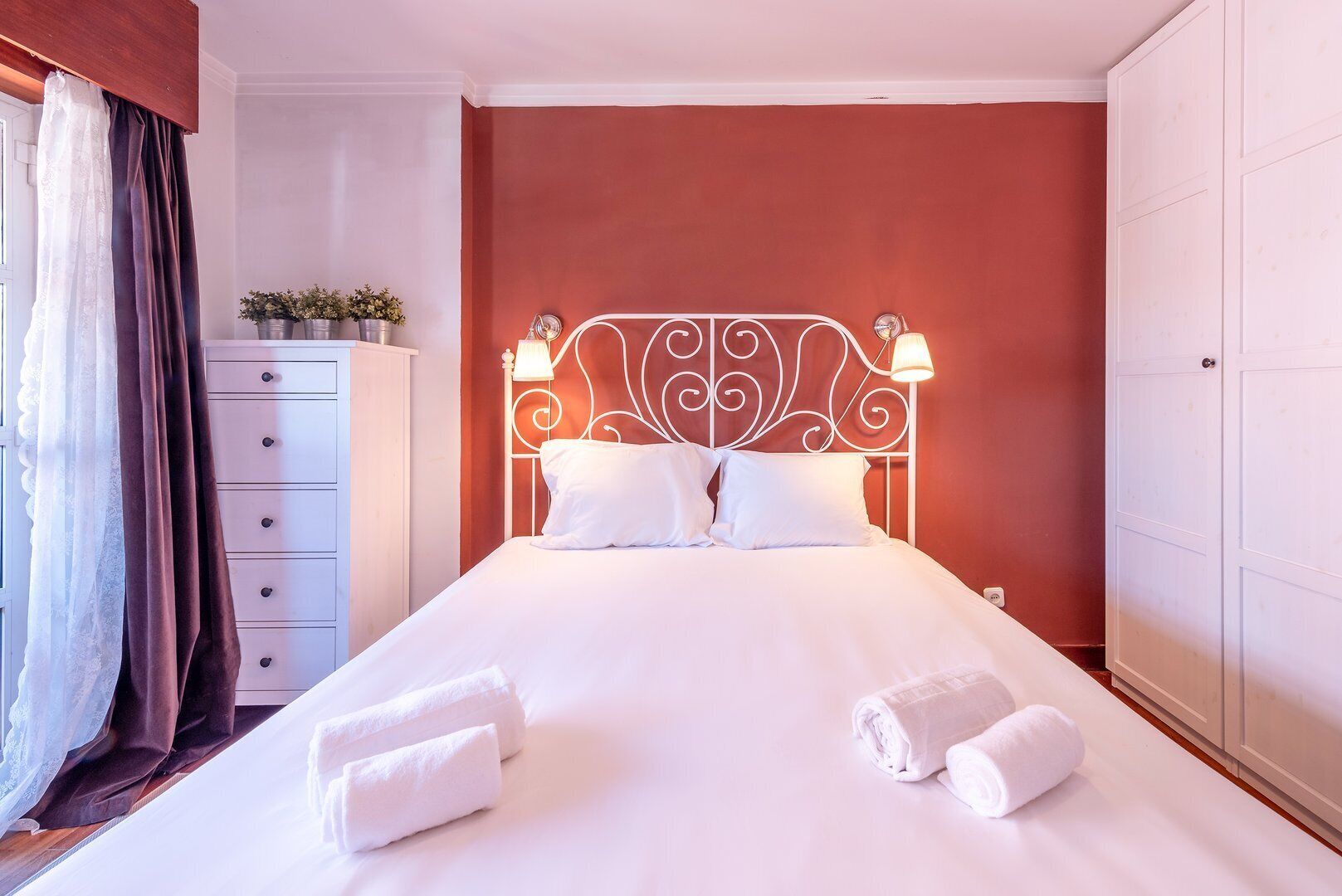 A neatly made bed with white linens and pillows, a white dresser with a mirror, and a red accent wall.