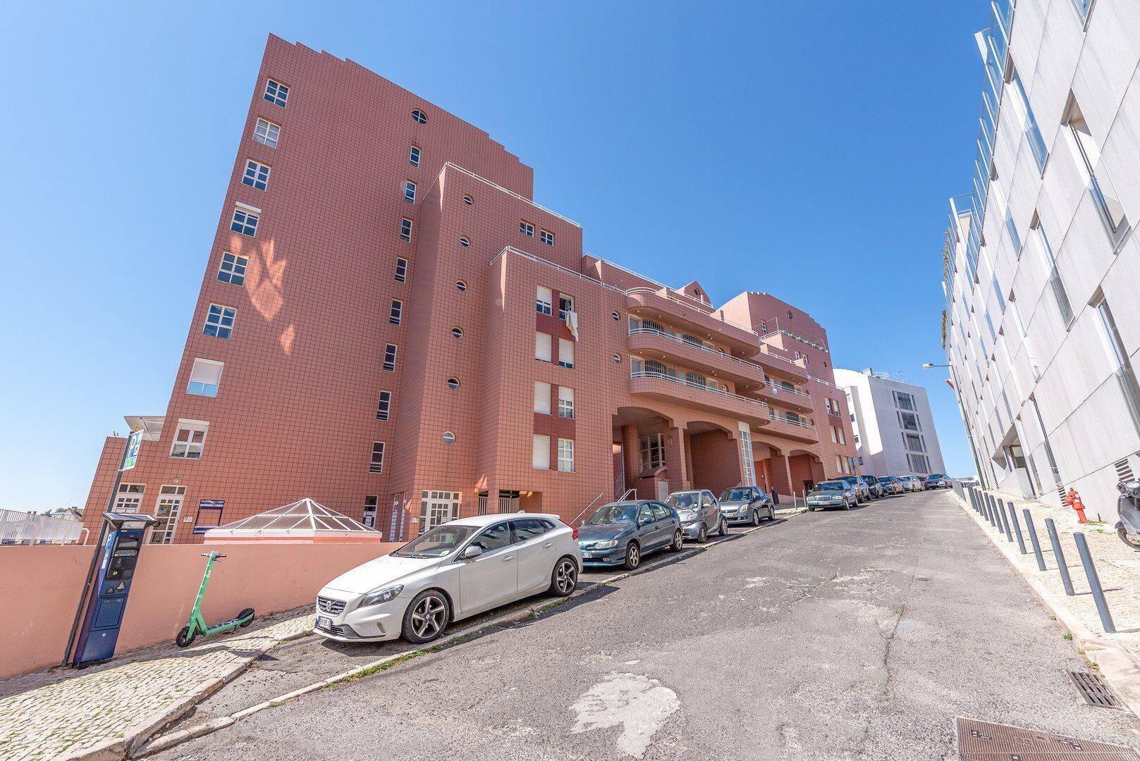 A multi-story building with a red brick facade, a parking area with several cars, and a clear blue sky.